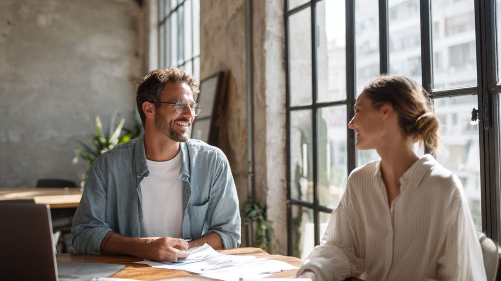 Adviseur en ondernemer in gesprek aan tafel met documenten, glimlachend in een lichte kantoorruimte